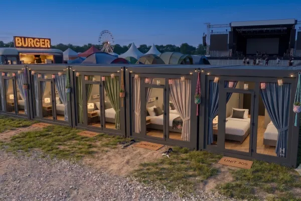 Hotel-Container - Mobile Unterkünfte für Ihr Event - Foto Vier nebeneinander aufgereihte, anthrazitfarbene Hotel-Container mit großen Glasfronten und gemütlich eingerichteten Zimmern auf einem Festivalgelände bei Dämmerung, im Hintergrund sind ein Riesenrad, Zelte und eine Konzertbühne zu sehen.