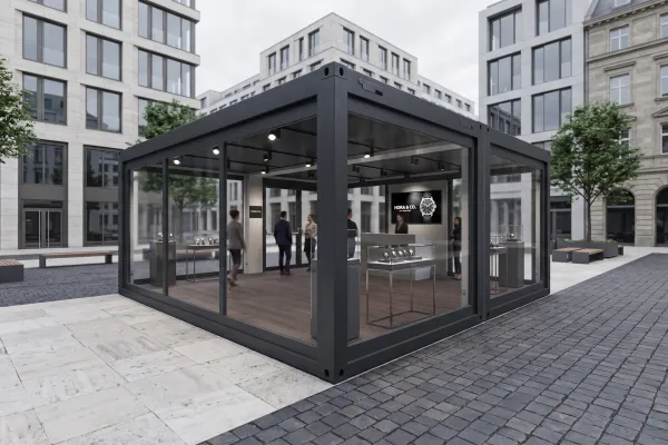 Luxury Showroom „Hora“ für Uhren & Schmuck - Foto Außenansicht eines modernen, schwarz gerahmten Glascontainers (Showroom) auf einem gepflasterten Stadtplatz, in dem Uhren und Schmuck in Vitrinen präsentiert werden.